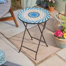 Round folding side table with a blue and yellow mosaic top on a patio surrounded by potted flowers.
