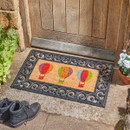 Small coir doormat with three colourful hot air balloons, set inside an ornate black rubber tray in front of a wooden door.