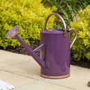 A tall glossy purple metal watering can with a rose sprinkler spout and copper-coloured handles, standing on a patio beside green shrubs.
