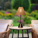 Rattan table lamp with a black metal base, lit and styled on an outdoor table between patio armchairs at night.