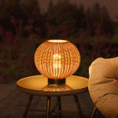 Round rattan table lamp glowing warm white on a glass-topped outdoor side table at night.