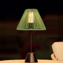 Sage green woven table lamp with a black base, glowing warmly on an outdoor side table.