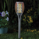 Slate-grey solar torch light glowing with a flame effect, standing in a garden at night beside flowers.