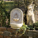 Ornate stone fountain with intricate patterns, surrounded by lush greenery, sits on a brick ledge beside a weathered classical statue in a garden.