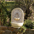Decorative stone fountain with intricate patterns, mounted on a brick wall, surrounded by lush greenery and bathed in warm sunlight.
