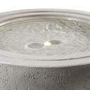 A close-up of a textured, gray concrete bowl fountain filled with water, showing gentle ripples and a soft light reflection on the surface. Calm and serene ambiance.