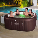 Four adults relaxing in a brown Lay-Z-Spa Maldives inflatable hot tub beside an outdoor swimming pool at night.