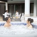 Man and woman relaxing in a bubbling Lay-Z-Spa inflatable hot tub with a modern house in the background.