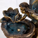 Angled close-up of the fairy and flower spout on a bronze-effect water feature, showing water flowing into the bowl with a warm LED light beneath the surface.