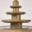 Close-up of a classic four-tier stone-effect garden fountain, with water cascading from the top finial into each rounded tier, against a white brick background.