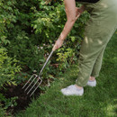 A person in green pants uses a garden fork to cultivate soil near lush green shrubs on a sunny day, conveying a sense of calm and care in gardening.