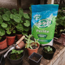 Bag of Gro-Sure Perlite sits on a wooden bench surrounded by potted plants, gardening tools, and pots. The setting conveys a calm, productive gardening environment.