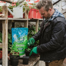 Man potting plants in a greenhouse, wearing a black jacket and green gloves. A Gro-Sure Perlite bag and red flower trays are on wooden shelves nearby.
