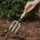 A hand uses a wooden-handled garden fork to till soil in front of a lush green bush. The scene conveys a sense of tranquility and gardening.
