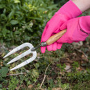 Hands wearing bright pink gardening gloves use a hand fork tool to tend to plants in a garden. The scene conveys an active, nurturing vibe.