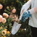 Person wearing gloves holds a gardening trowel near blooming peach dahlias and yellow flowers. The scene conveys a sense of tranquility and care.