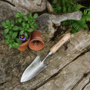 A small trowel with a wooden handle rests on weathered wood beside two terracotta pots, one empty and one with green plants and a purple flower.