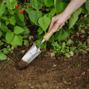 A hand uses a metal garden trowel to dig in rich soil near leafy green plants with visible red flowers, conveying a sense of gardening and growth.