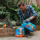 A man in a plaid shirt and gloves is pouring Resolva weedkiller from a large blue container into a pump sprayer. He's kneeling by a flowerbed with blooming flowers, and a brick border. The scene is calm and focused on gardening.