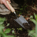 A hand using a wooden-handled trowel digs into rich, dark soil surrounded by green foliage, conveying a sense of gardening and nature.