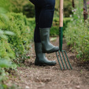 A person stands in an earthy garden path, wearing green boots and dark pants, holding a garden fork. Green foliage lines the path under soft daylight.
