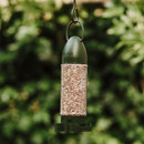 A green bird feeder filled with seeds hangs against a blurred leafy background, conveying a sense of tranquillity and inviting nature.