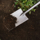 A shiny metal shovel lifting dark, rich soil in a garden, with green plants in the background. The tone conveys a sense of growth and gardening.