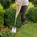 A person in a white shirt and green pants uses a spade to tend a garden with green bushes and blooming orange flowers, conveying a peaceful, gardening vibe.
