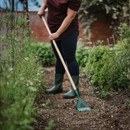 Person in jeans and boots using a hoe in a lush garden. The scene conveys calmness and concentration amidst green plants and a brick wall background.