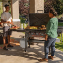 Two people cooking outdoors on a Blackstone 4-burner propane griddle on a patio.