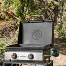 Blackstone tabletop griddle with hood open on camping table, two adjustable heat control knobs and rear-mounted green propane bottle in wooded outdoor setting.