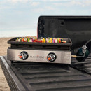 Blackstone portable gas griddle cooking vegetable skewers on a truck tailgate at the beach, stainless steel front panel with two heat control knobs and green propane bottle attached.