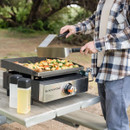 Person cooking outdoors on a Blackstone tabletop griddle with spatulas and oil bottles on a picnic table