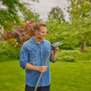 A man in a denim shirt stands in a lush garden, holding a gardening tool. He appears focused and contemplative with vibrant greenery around.