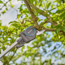 Gardening shears from the Gardena brand are cutting a tree branch against a backdrop of green leaves and a bright blue sky.