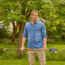 Man in denim shirt and beige pants holds gardening tool in lush, green garden. He looks thoughtfully into the distance, surrounded by trees.