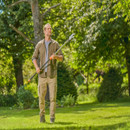 A man in an outdoor garden holds a pruning tool, gazing upwards at tree branches. He stands on a sunlit lawn, surrounded by lush greenery.