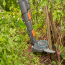 Gardening tool cutting thick branches among lush green foliage and stems. The tool has a sleek gray design with orange accents, conveying efficiency.