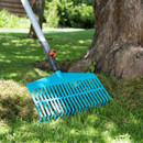 A blue garden rake leans against a tree trunk on green grass. Sunlight casts shadows, creating a peaceful, outdoor gardening scene.