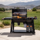 Traeger Woodridge Pro pellet grill cooking outdoors on a patio, lid open with brisket, ribs, vegetables and corn on the grill, digital controller lit and storage bins below.