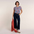 Woman smiling in a cream floral print T-shirt with blue leaves and red flowers, paired with dark indigo wide-leg jeans and trainers, holding a coral jacket.