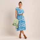 Woman wearing a blue and white floral midi dress with short flutter sleeves and a brown belt, styled with tan ankle boots and holding a light green jacket, against a neutral studio background.