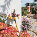 Close-up of a garden hose splitter attached to a wall among colourful flowers. A person in the background waters plants under a sunny sky.