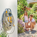 A garden hose with yellow nozzles hangs neatly on a wall. Nearby, a woman and a young girl admire vibrant purple flowers in a sunny garden, creating a joyful scene.