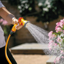 A person waters pink flowers using a yellow and red garden hose nozzle, spraying a gentle mist. The background is a blurred garden scene.