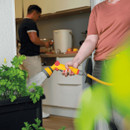 A person in a salmon shirt waters a vibrant potted plant with a yellow hose on a patio. Inside, a person in black prepares food, adding a domestic feel.