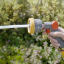 A person holds a Hozelock garden hose sprayer, with water spraying out, set against a vibrant, blurred green garden background in sunlight.
