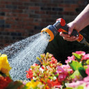 A hand holds a garden hose nozzle, spraying water onto vibrant flowers, including yellow and pink blooms, against a blurred brick wall background.