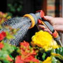 A person waters colourful blossoms, including red and yellow flowers, with a hose spray nozzle, creating a refreshing, vibrant garden scene.