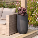Tall ribbed outdoor planter in dark grey, displayed on a patio beside a wicker garden sofa and wooden post, planted with purple foliage.
